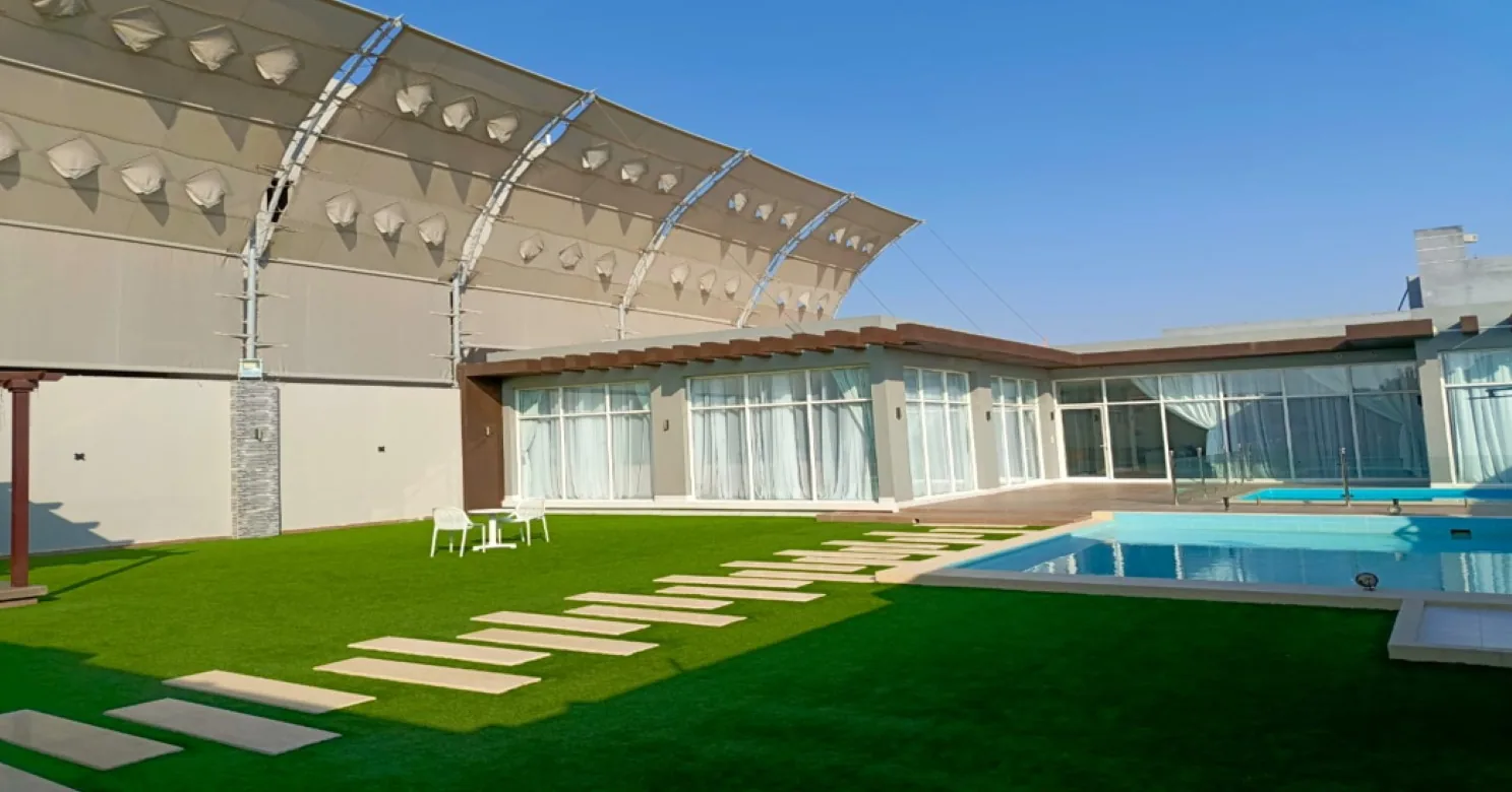 Finished exterior with a tensioned fabric canopy on steel arches, glazed pavilion, green turf with stepping slabs and a pool at right.