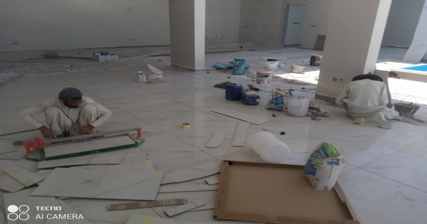 Two workers tiling an indoor floor with large white marble-look tiles, one at a manual tile cutter, amid tools and adhesive bags.