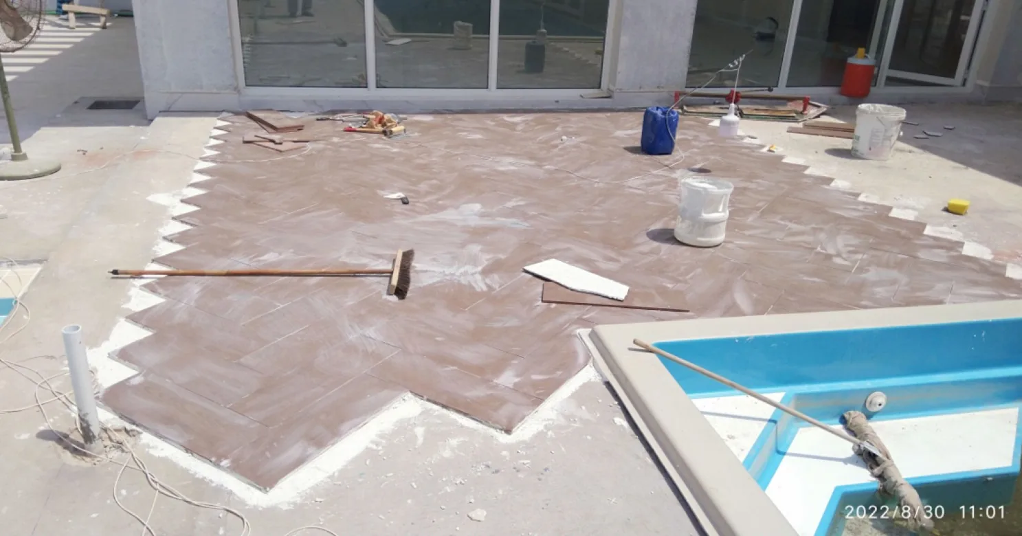 Brown rectangular tiles being laid in herringbone pattern on a terrace beside a pool, with cement dust, a broom and buckets.