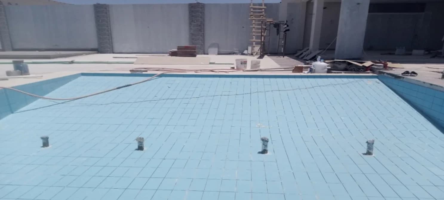 Empty tiled pool with a hose trailing along its edge; beyond it a deck with materials, a ladder and stone-clad wall pilasters.
