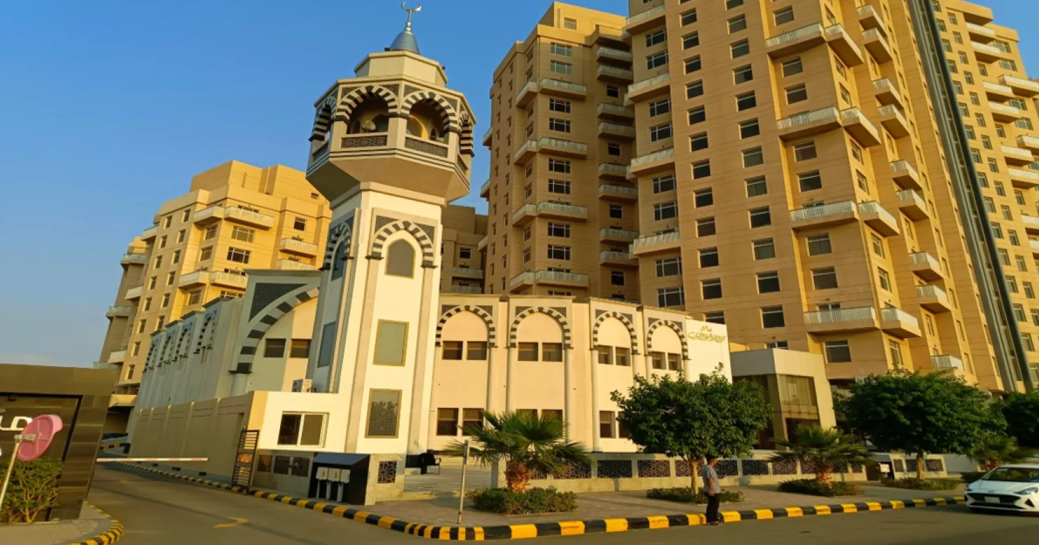Mosque minaret with black-and-white banded arches and a cream mosque frontage, set against ochre residential towers in warm evening light.