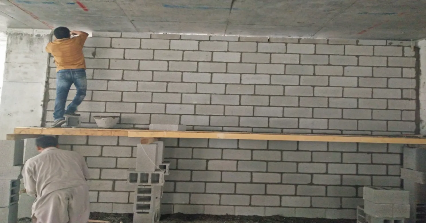 Two workers building a concrete-block wall, one standing on a timber plank resting on stacked blocks to reach the upper courses.