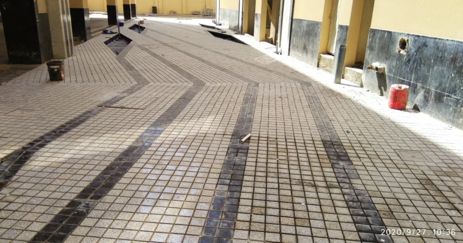 Granite mosaic paving with dark inlay bands along a colonnaded walkway, dark stone wall dado and open drainage channels not yet closed.
