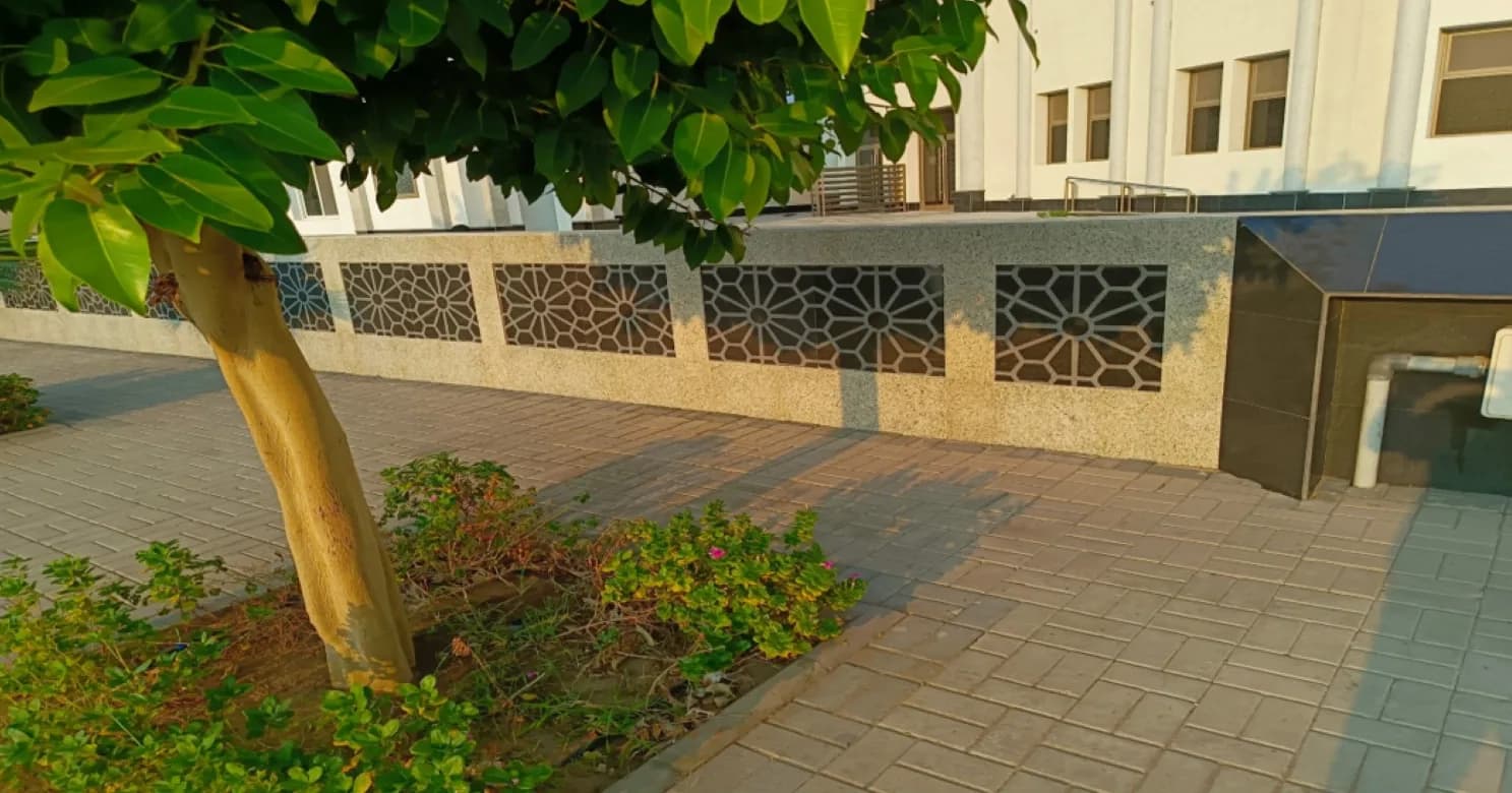 Leafy tree beside a low speckled-stone wall with geometric star-pattern openings, above interlock paving in warm evening light.