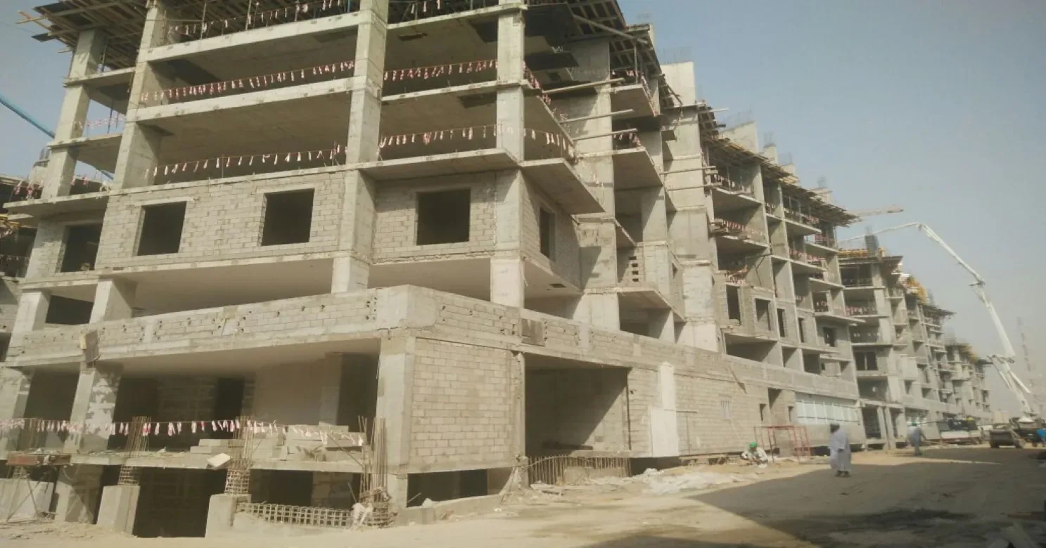 Concrete-frame residential blocks under construction, with blockwork infill, open window openings, marker bunting and a pump boom at right.