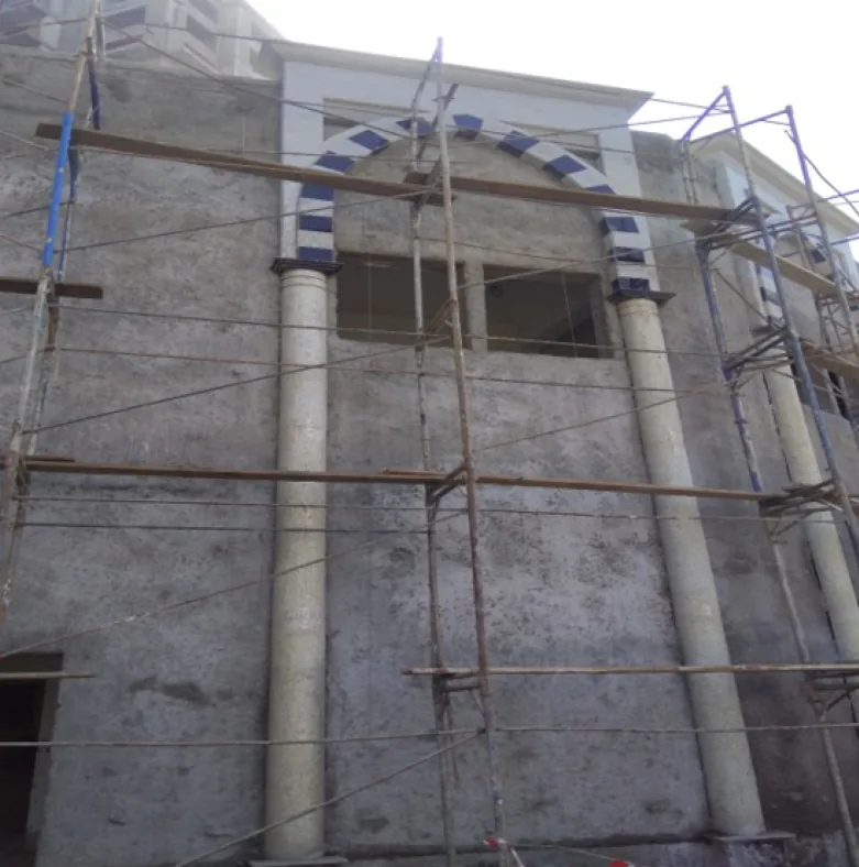 Scaffolded rough-rendered facade with two pale stone columns carrying an arch banded in dark blue and white above two window openings.