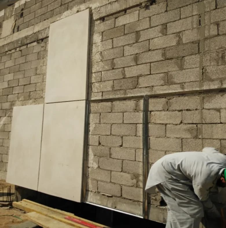 Worker fixing large pale stone-look panels to a blockwork wall on steel channels; three panels are up, the next bay is bare.