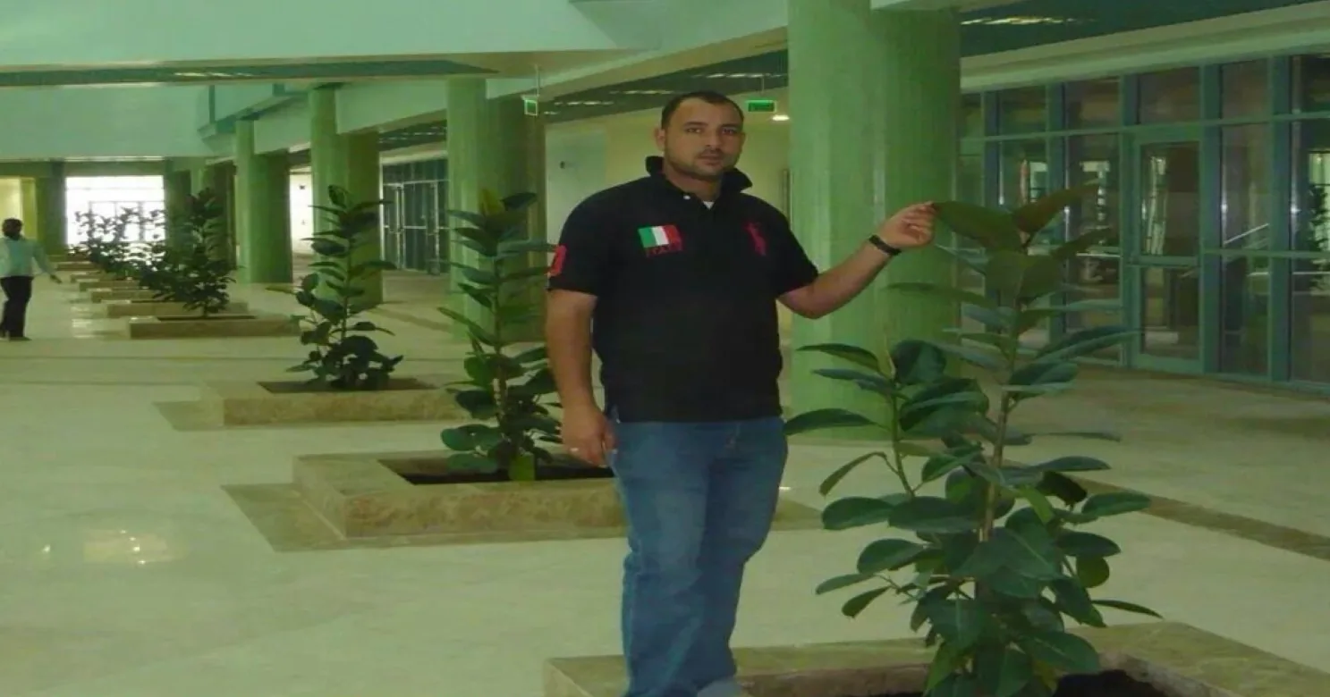 Atrium arcade with green columns and ficus plants in stone-edged planters; a man in a black polo shirt poses at centre.