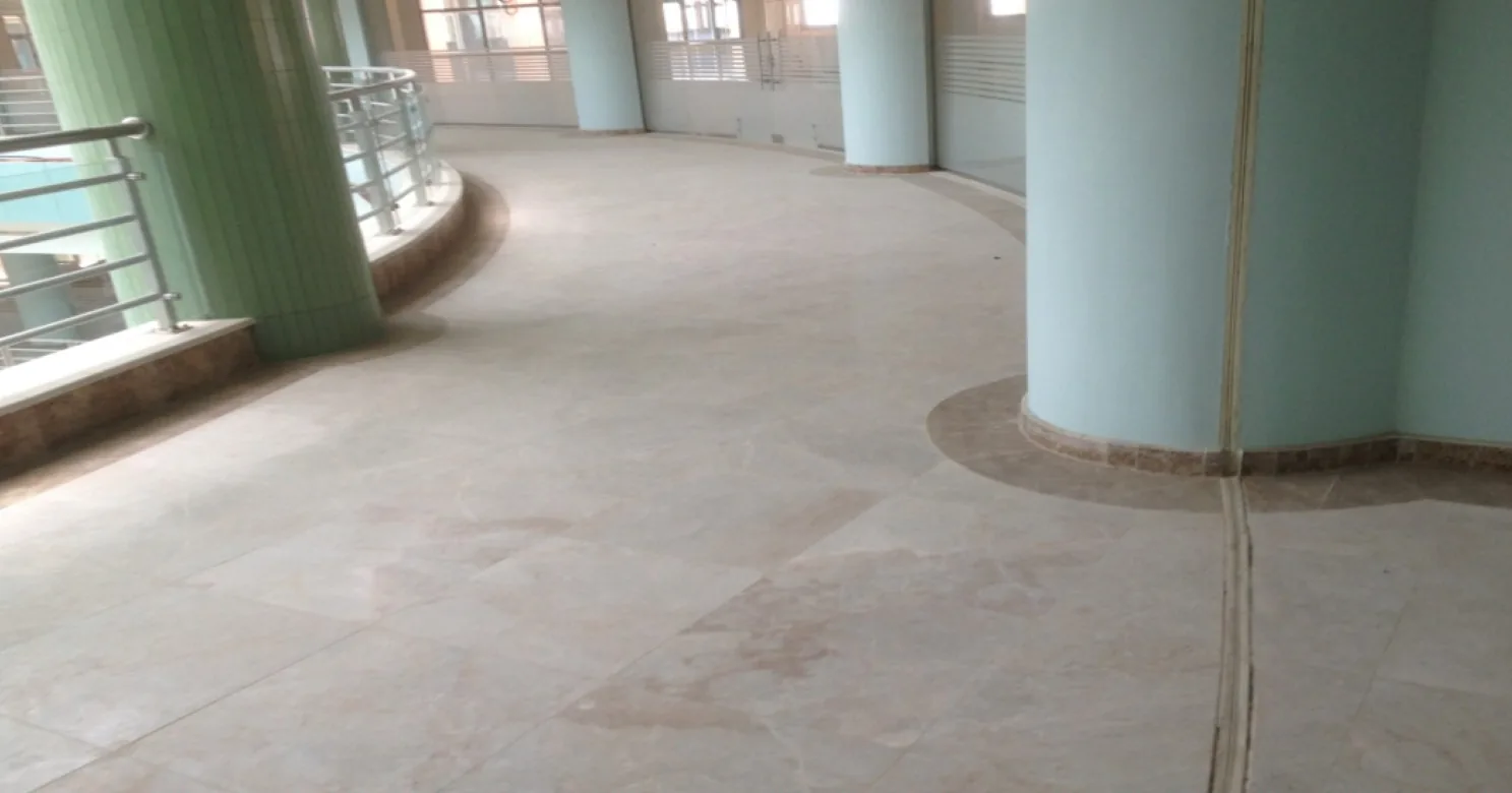 Curved gallery floor in large pale stone slabs around a green-tiled column and pale-blue columns, with a movement-joint strip and steel railings.