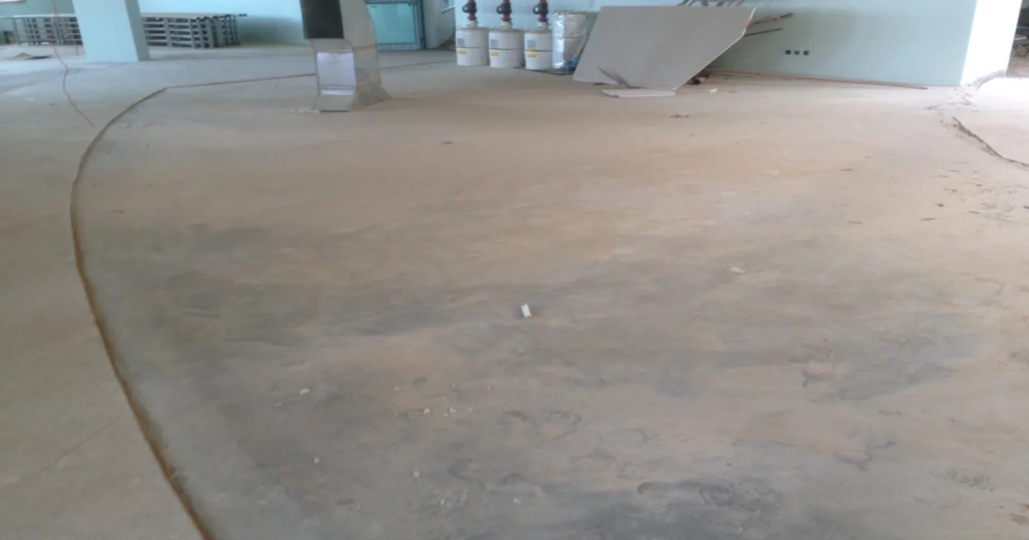 Bare, dusty curved floor still awaiting finishes, with a curved joint line, sheet-metal ducting and a board leaning at the back wall.