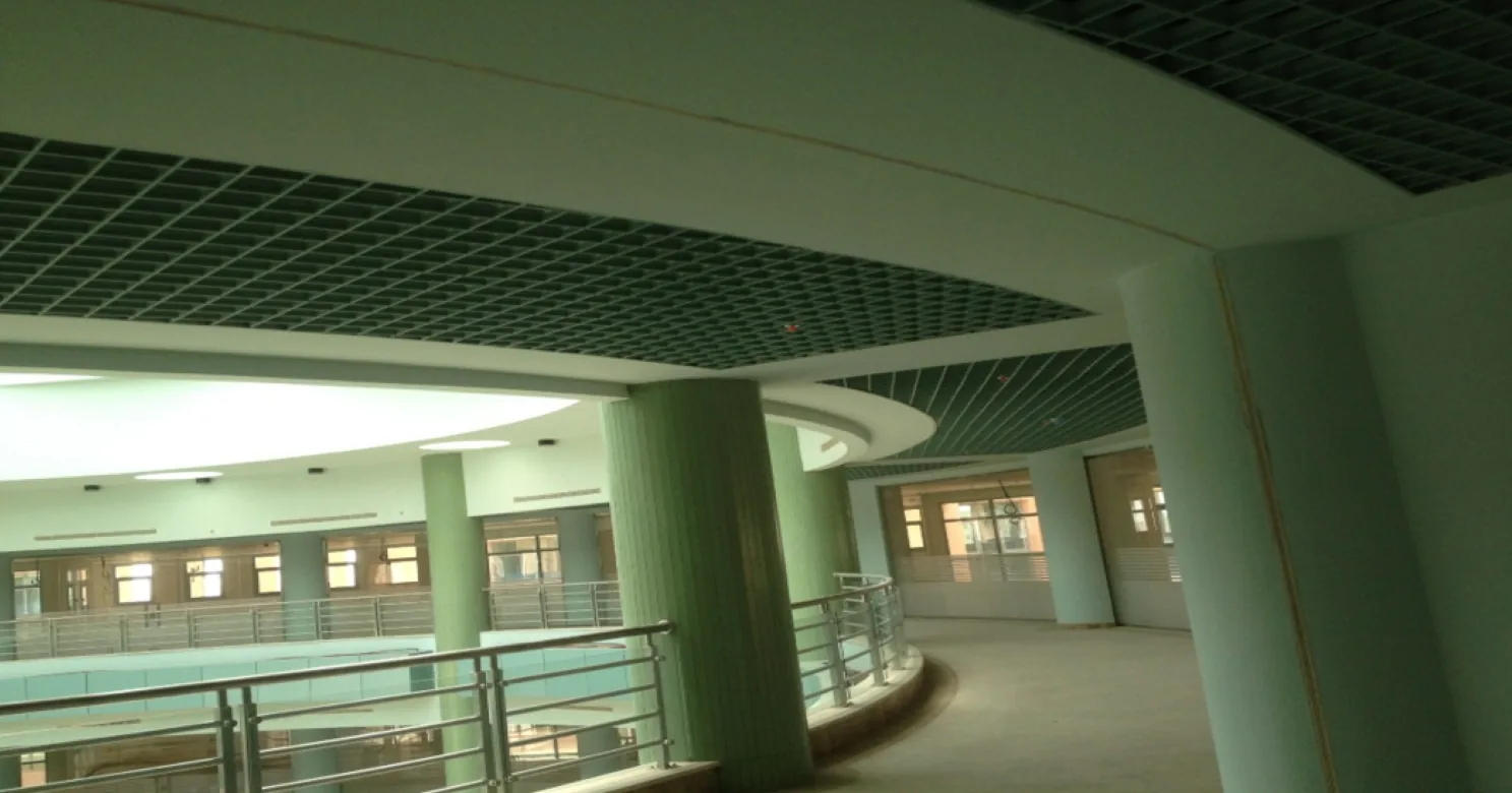 Curved gallery walkway under a dark open-cell grid ceiling, with white curved bulkheads, green columns, steel railings and a bare floor.