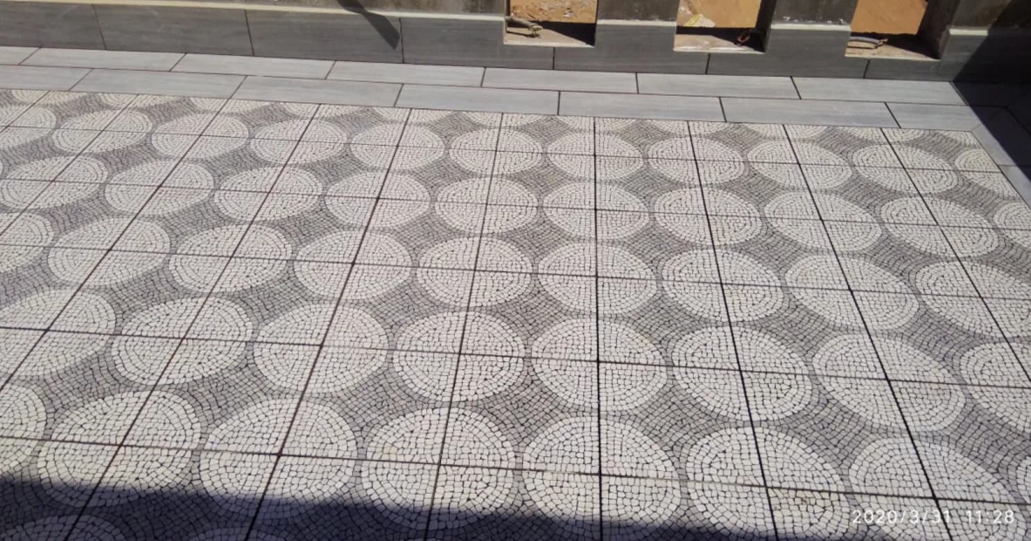 Terrace floor of round-motif mosaic tiles bordered by grey plank-look tiles beside a low wall with openings.
