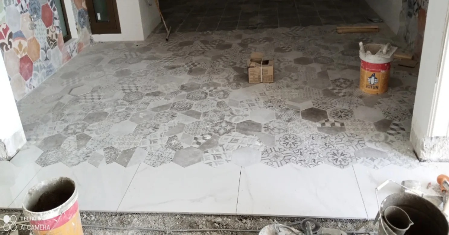 Floor tiling in progress at an entryway: patterned grey hexagon tiles meeting large white marble-look tiles, adhesive buckets in front.