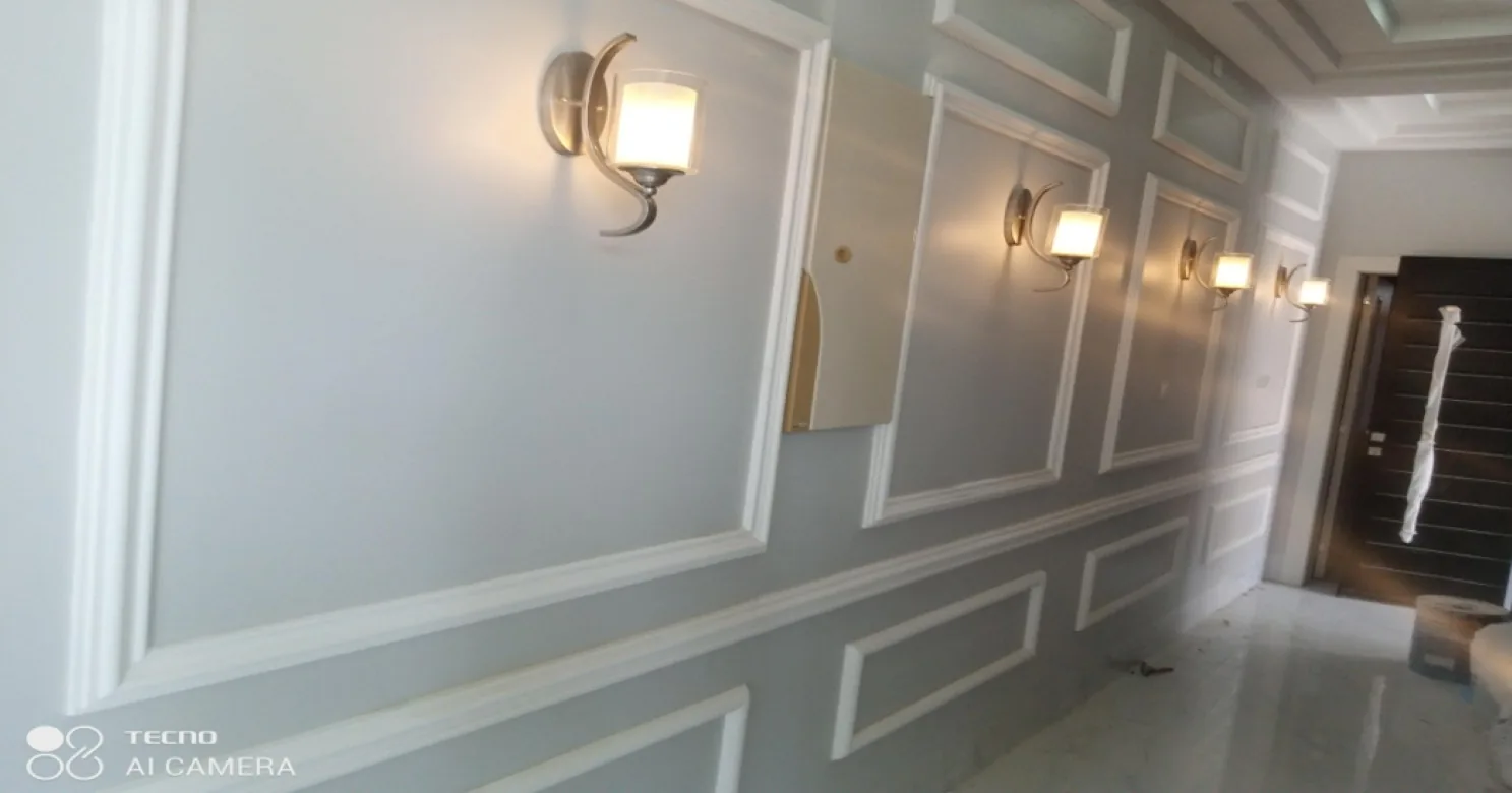 Corridor with grey walls framed by white mouldings, wall lights, a polished marble-look floor and a dark door at the end.