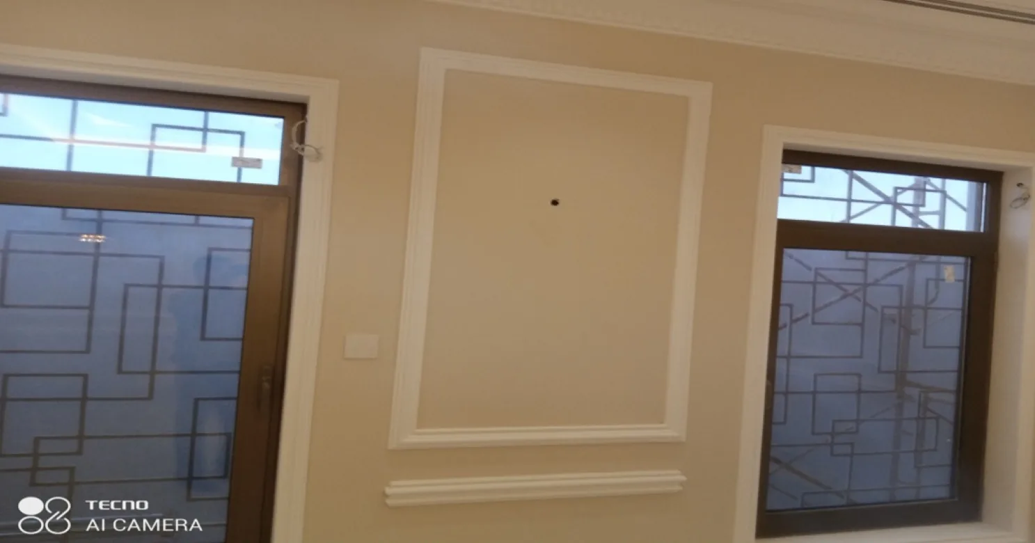 Cream room with a white moulded panel between two dark-framed aluminium windows with geometric grille patterns, scaffolding outside.