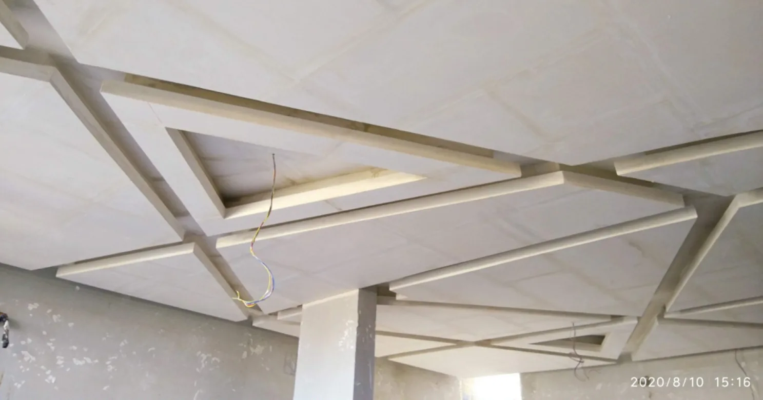 Unfinished stepped gypsum ceiling built as a grid of boards and beams, with wires hanging down and a column below.