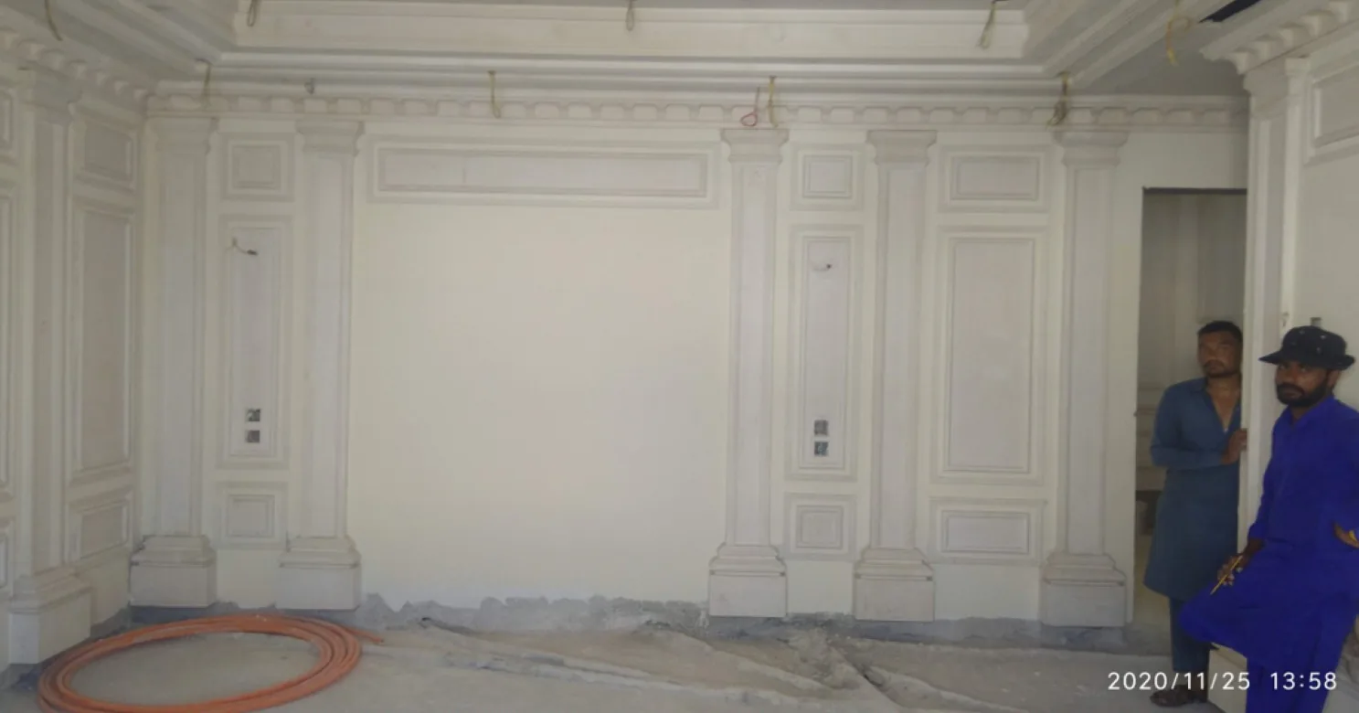 Unpainted classical gypsum wall with pilasters and panel mouldings, an orange conduit coil on the floor and two workers standing at right.