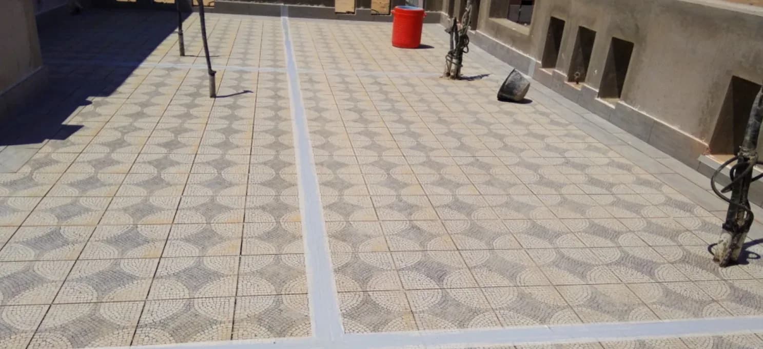 Roof terrace floored in circle-motif mosaic tiles crossed by wide sealed grey joint bands, with pipe risers and a red container.