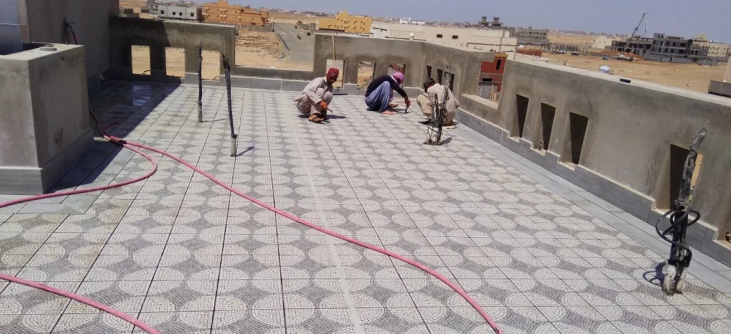 Three workers crouched on a roof terrace, laying or grouting circle-motif mosaic tiles, with a pink hose across the floor.