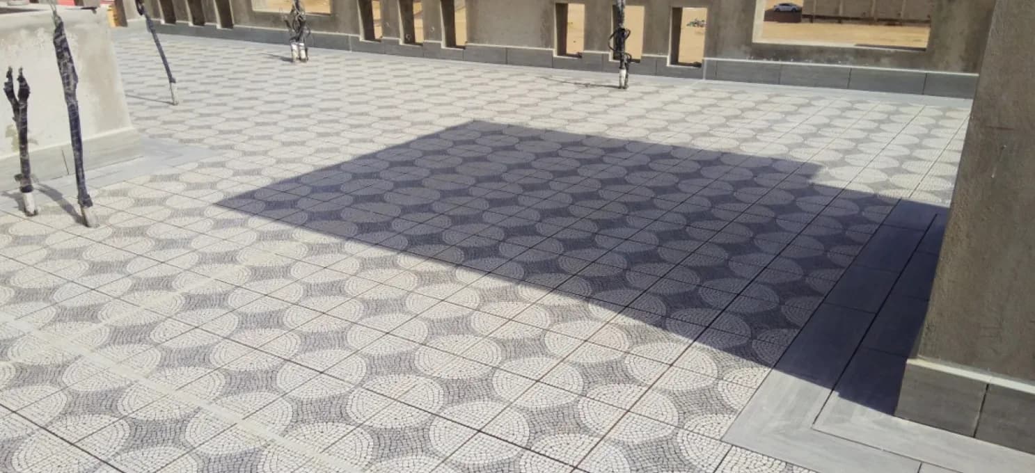 Roof terrace of circle-motif tiles with a plank-look border, a large cast shadow and pipe risers along a parapet with openings.