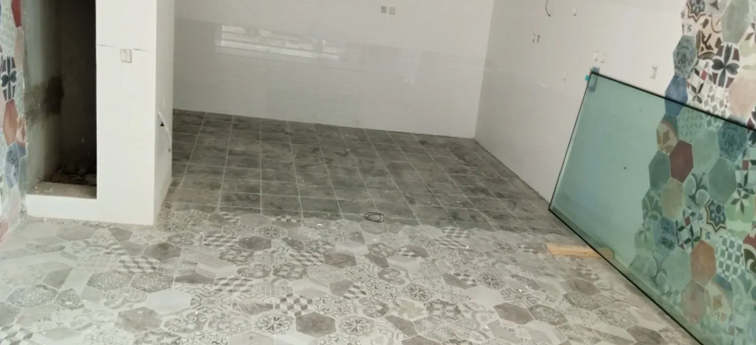 Room with white glossy wall tiles, grey square floor tiles turning to patterned hexagons, and a glass panel leaning at right.