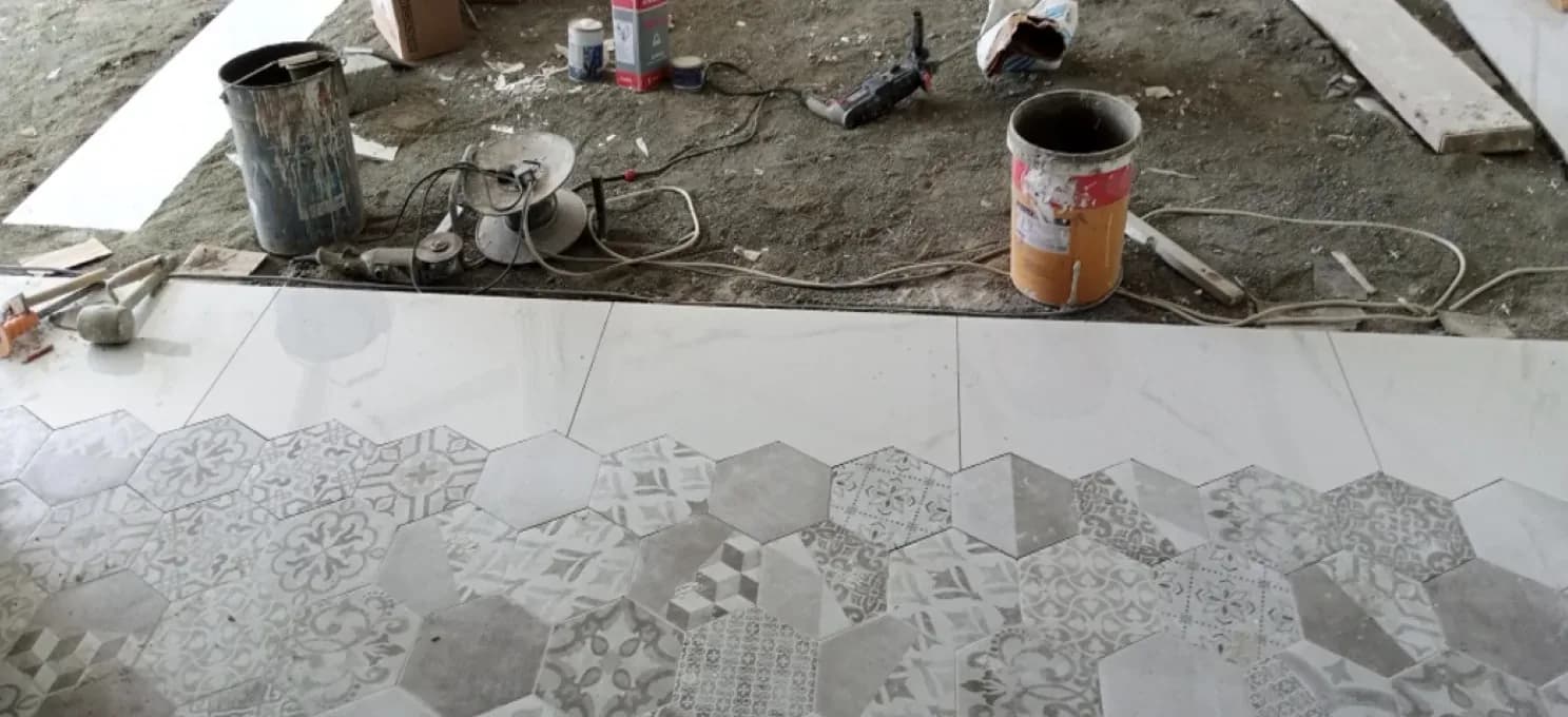 Large white marble-look tiles meeting patterned hexagon tiles on a floor being laid, with tools and buckets on the bare ground beyond.