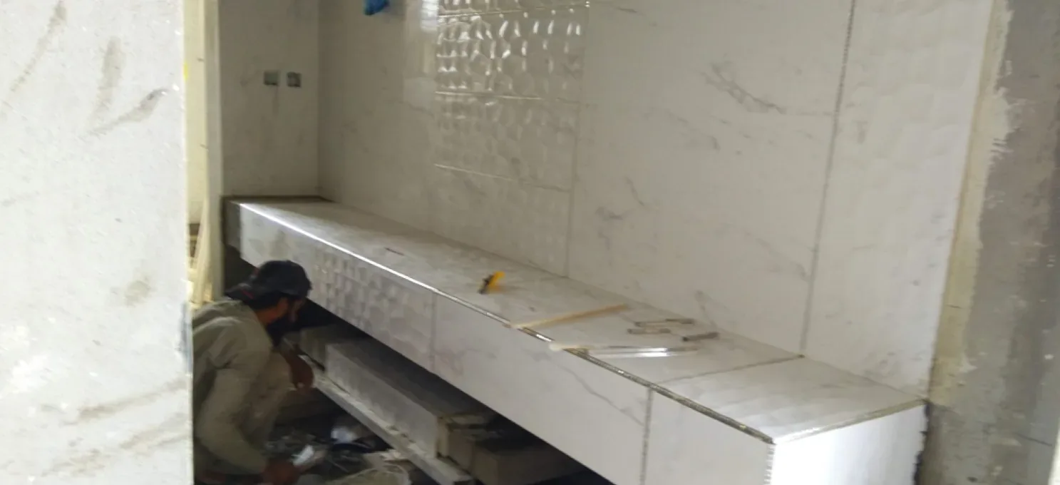 Worker crouched under a counter being clad in white marble-look tiles, with a textured tile panel on the wall behind.