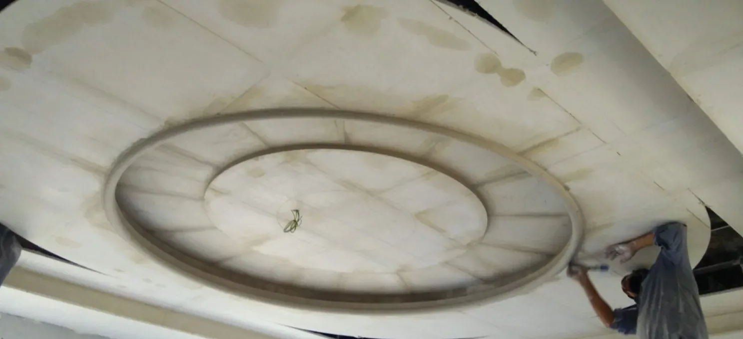 Worker smoothing the edge of a curved oval ring in a stained, unfinished gypsum ceiling, with board joints visible.