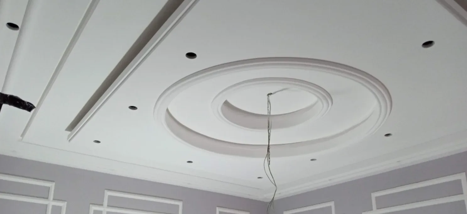 Painted gypsum ceiling with a concentric ring rose, recessed light openings and a hanging cable, above lilac-grey walls with white frame mouldings.