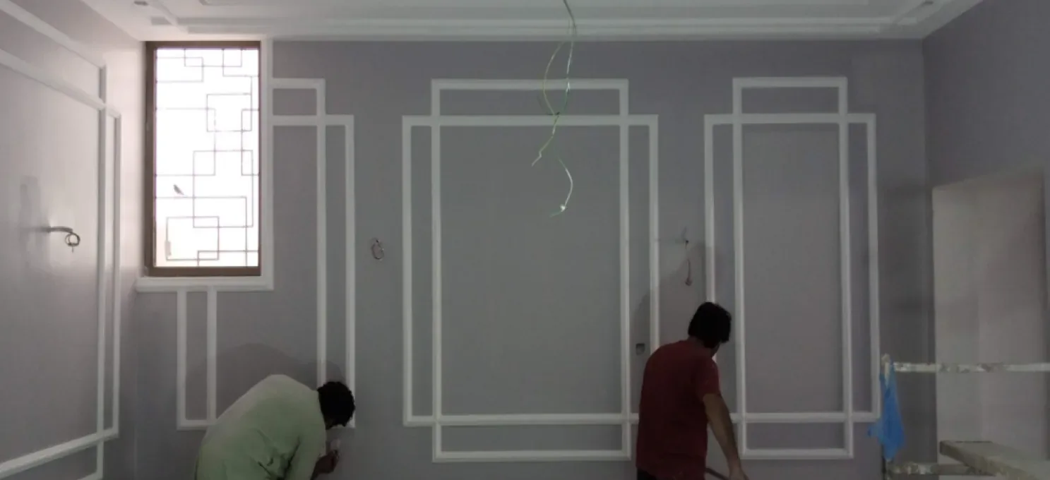 Lilac-grey wall with white geometric frame mouldings and a tall grille window, with two workers at work near its base.