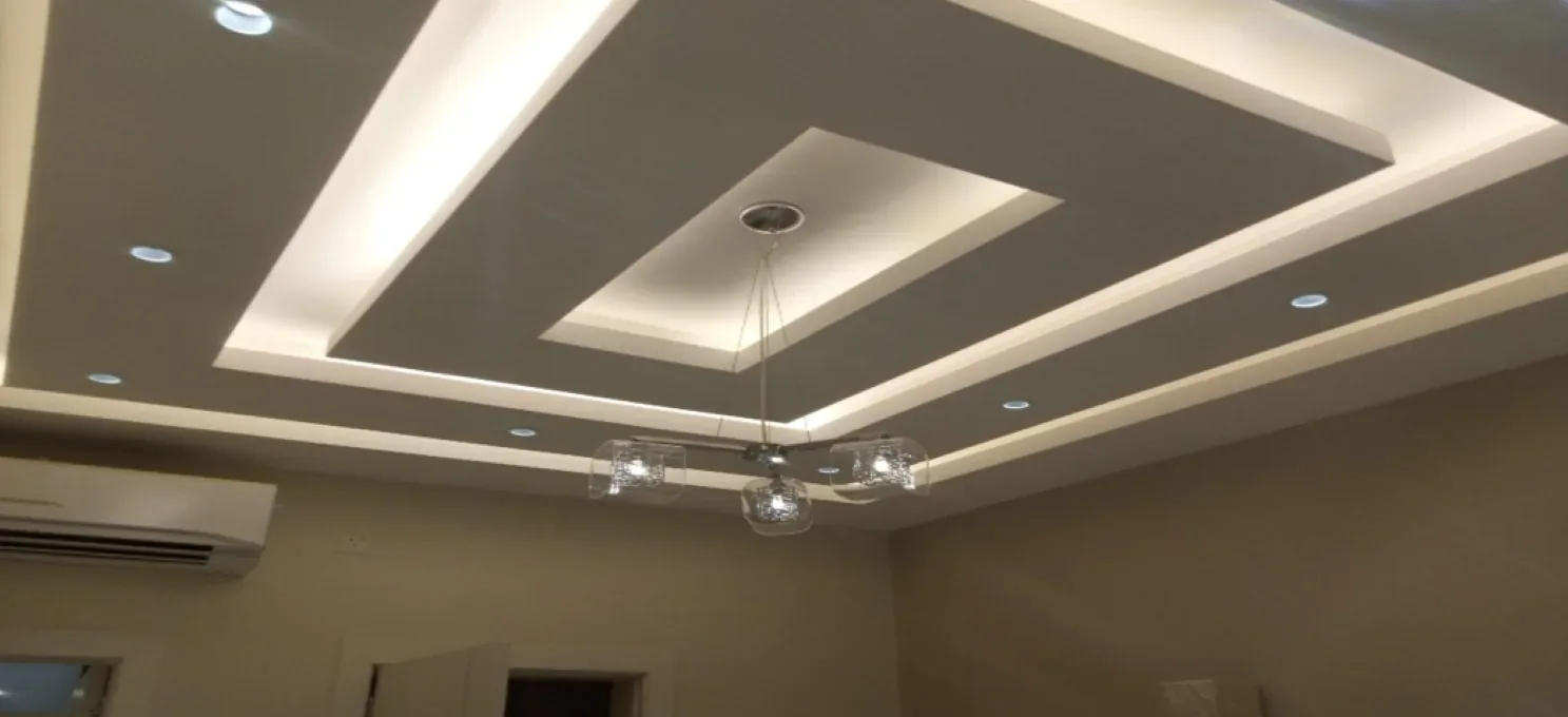 Cove-lit stepped ceiling with recessed downlights and a three-shade glass pendant, above beige walls and a wall-mounted air conditioner.