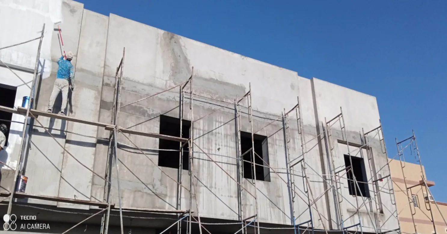 Worker on a scaffold plank rolling a white coat onto a bare concrete facade with window openings, scaffold towers along the front.