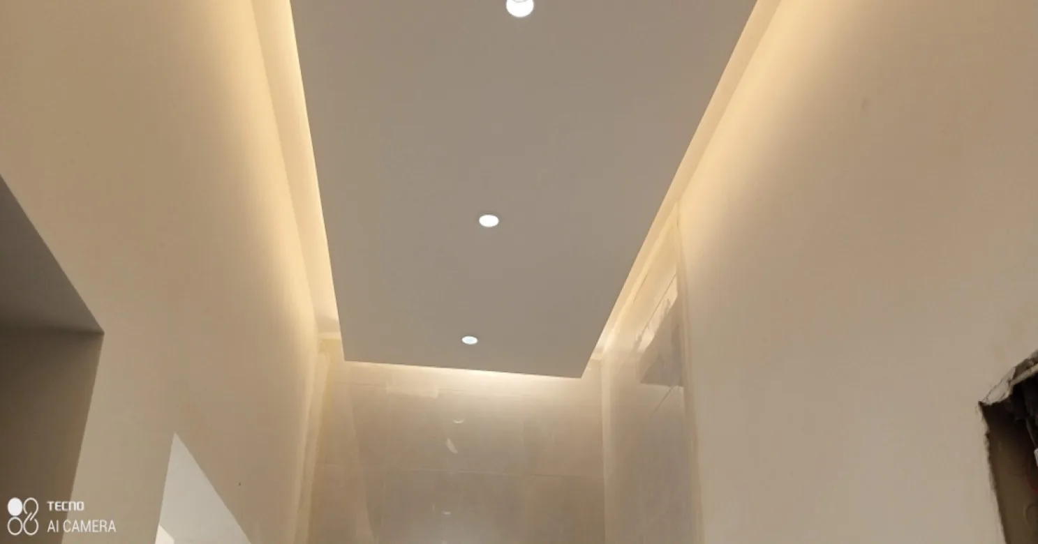 Narrow hall with a rectangular recessed ceiling panel, warm concealed cove lighting and three downlights above cream walls.