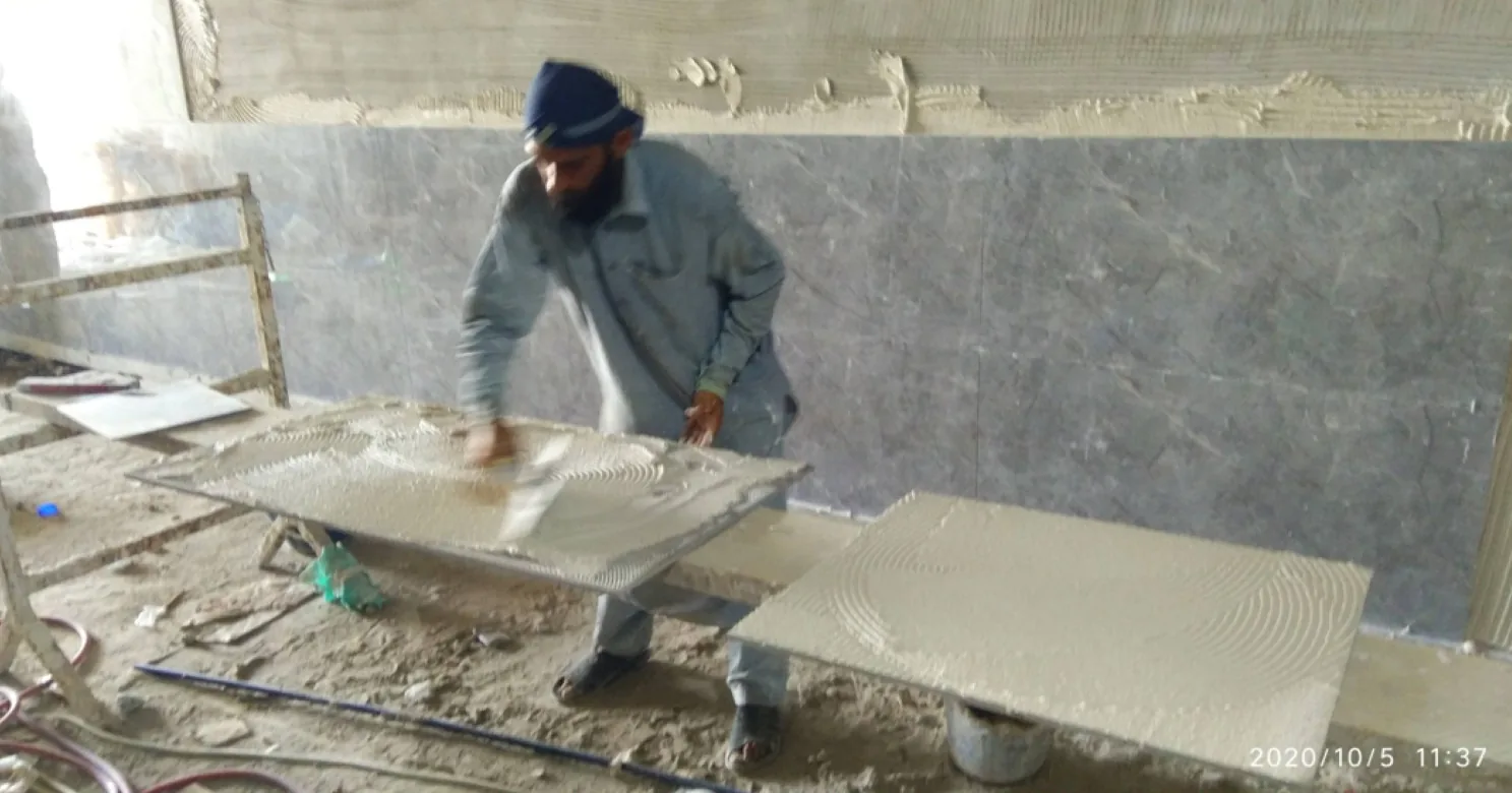 Worker spreading adhesive on a large slab, another slab combed with adhesive beside it, in front of grey veined wall cladding.