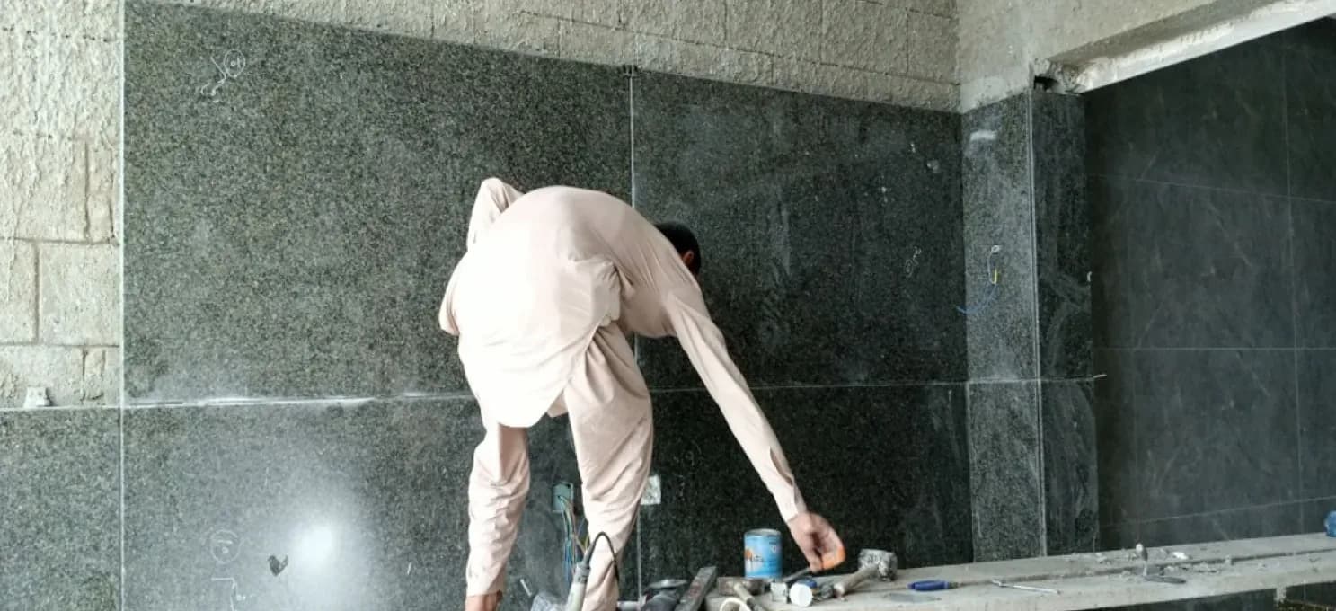 Worker bending over a ledge to fix dark granite wall slabs, with a hammer, a tin and tools beside him.