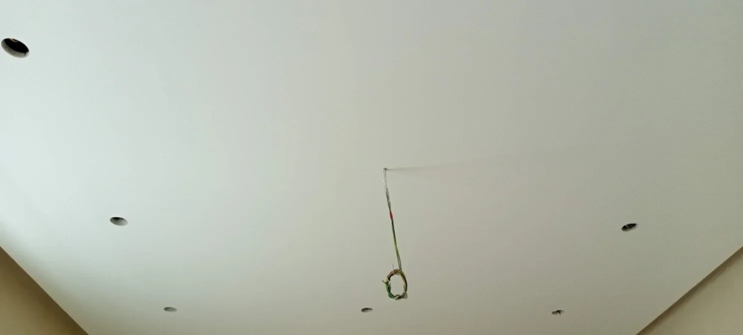 Smooth white ceiling showing six empty downlight holes and a wire coil at the centre drop point; cream walls at the edges.