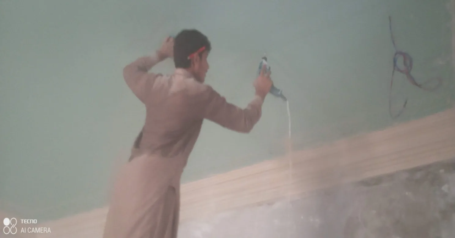 Worker in a thobe spraying material onto a green-tinted wall with a handheld tool in a dusty interior; the frame is blurred.
