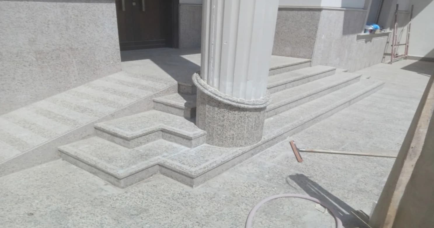 Granite entrance steps around a fluted column on a granite base, with wall-base cladding, a broom and a hose on the paving.