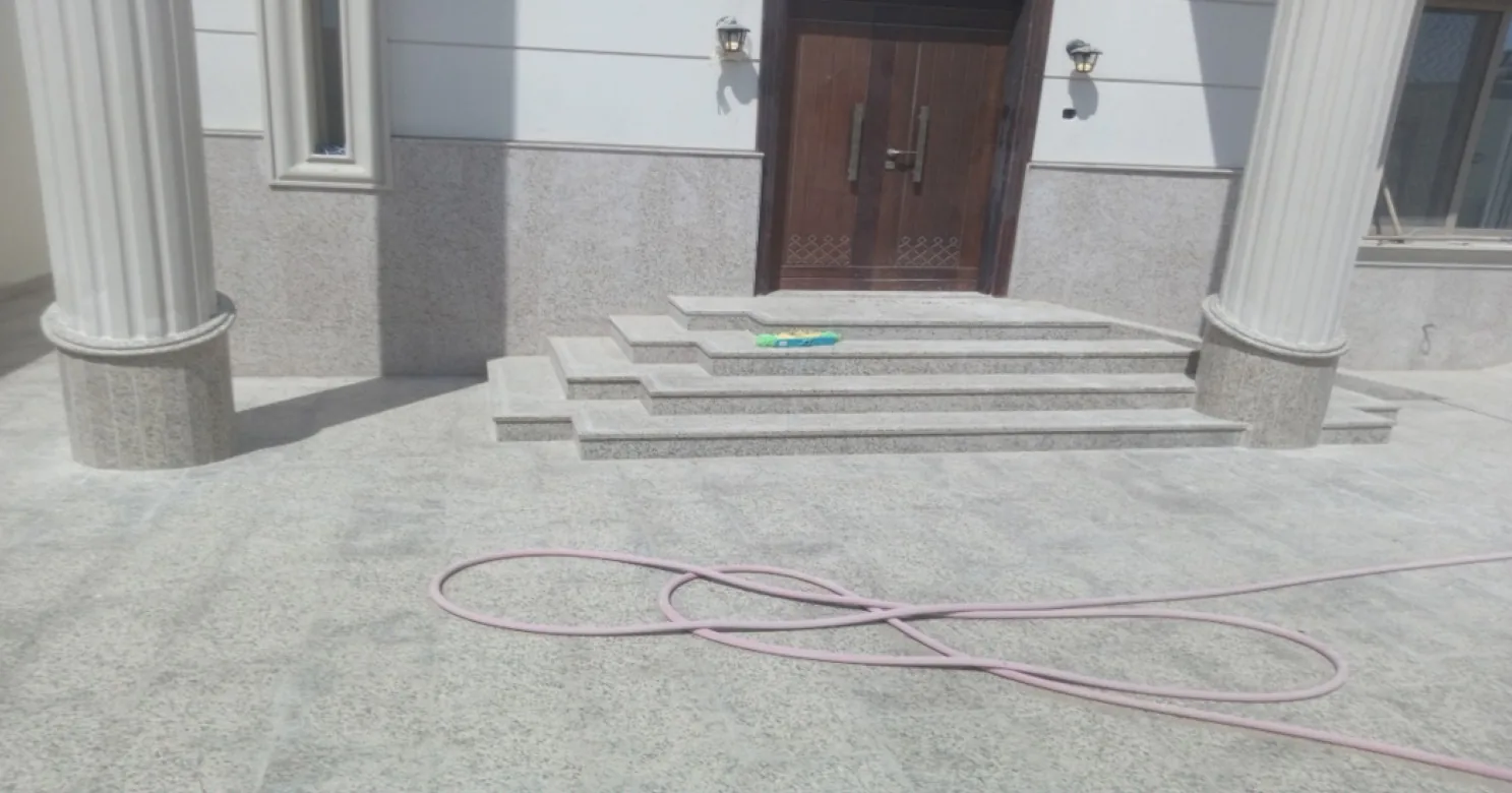 Frontal view of granite entrance steps up to a wooden double door between two fluted columns, a pink hose in the foreground.