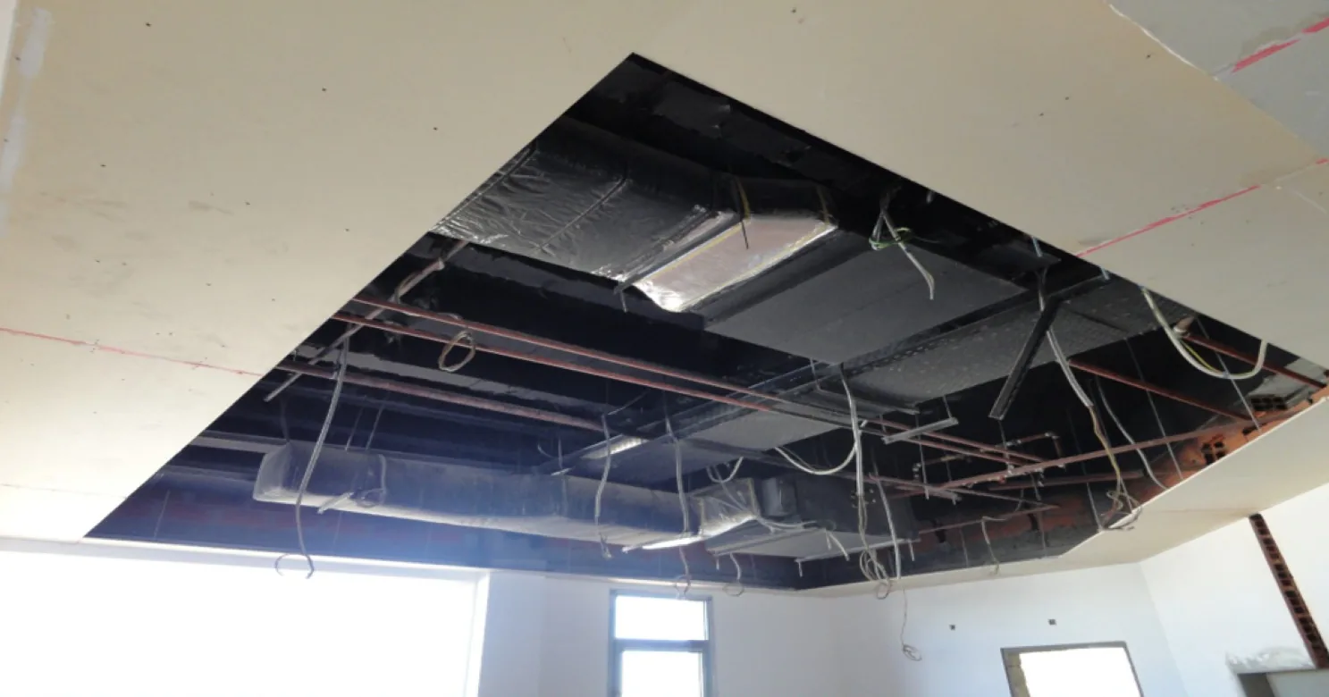Plasterboard ceiling with a large opening exposing insulated ducts, red pipes, a cable tray and hanging wires above the room.
