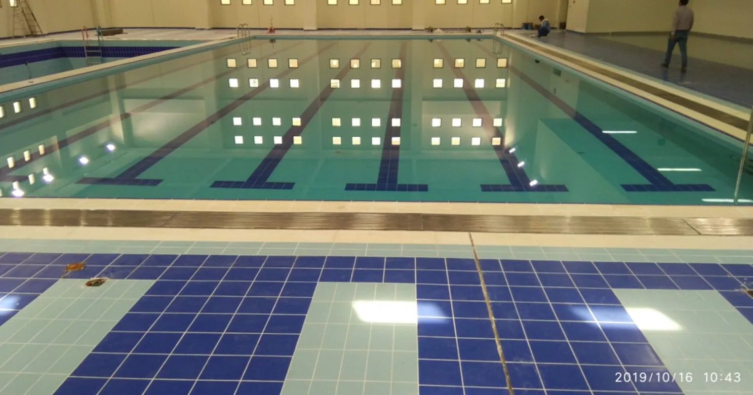Water-filled indoor pool with dark-blue lane lines on its floor and a dark- and pale-blue tiled deck in the foreground.