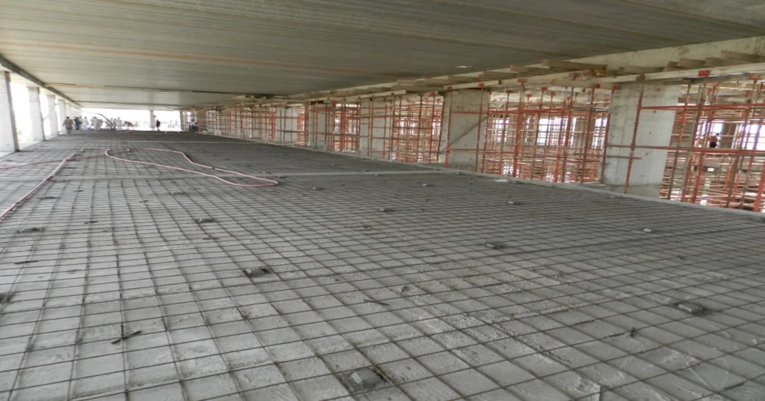 Wire reinforcement mesh laid across a wide slab under a steel-deck soffit, with orange props and scaffolding along the far edge.