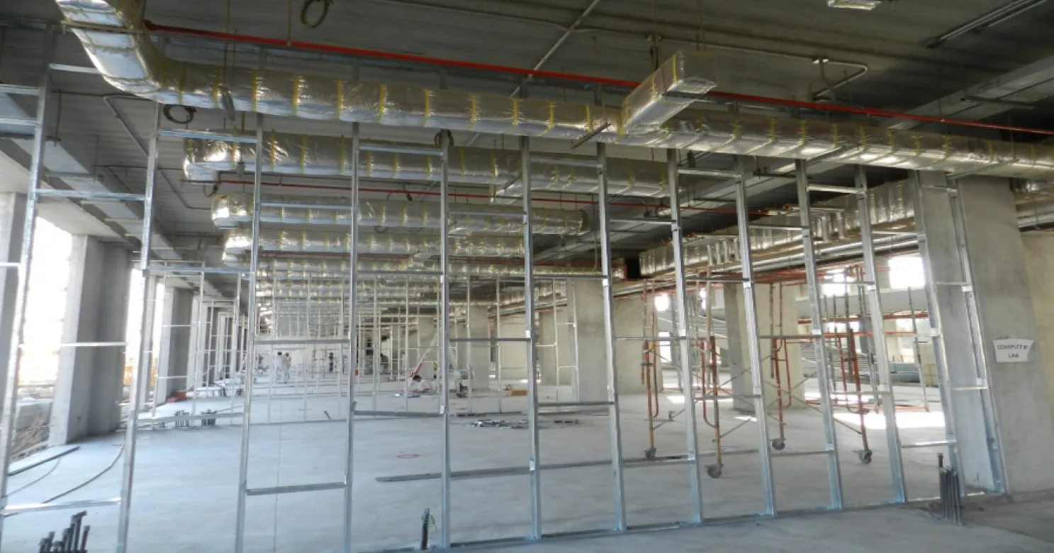 Galvanised steel-stud partition framing beneath foil-wrapped ductwork and red pipes, with rolling scaffold towers behind.
