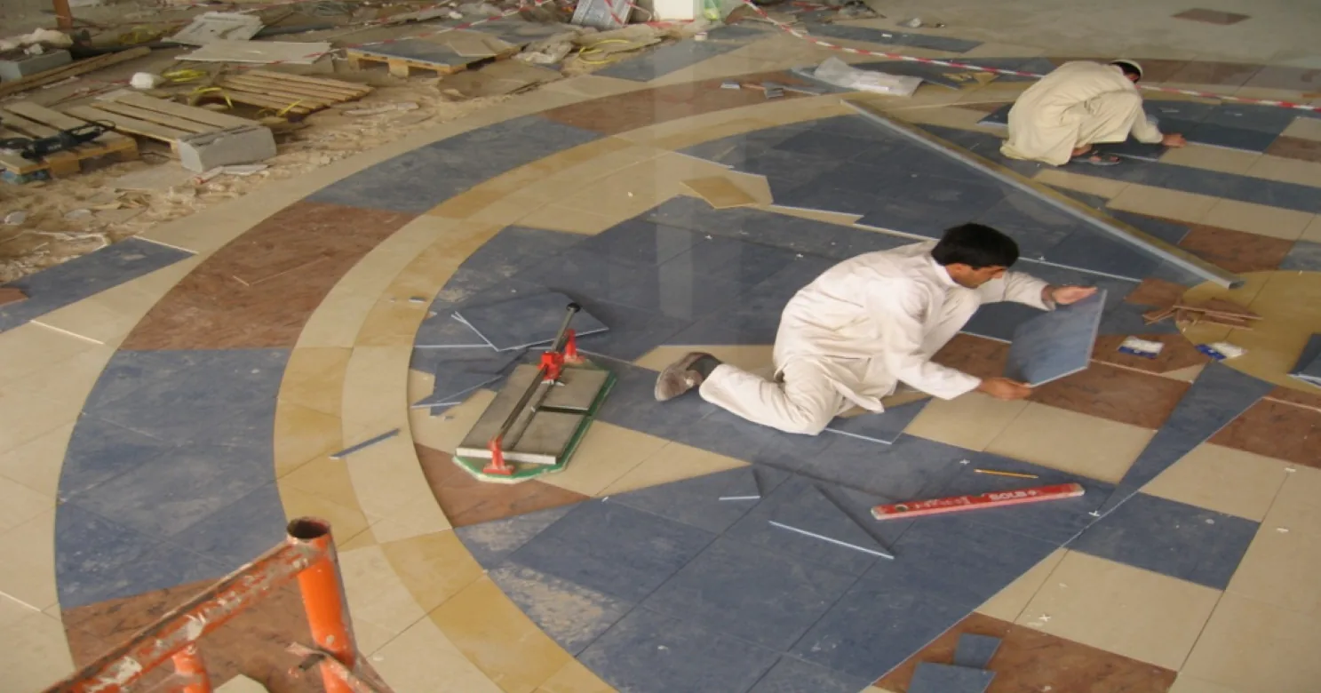 Two installers kneel on the medallion floor, laying blue, tan and terracotta tile segments in concentric arcs beside a hand tile cutter