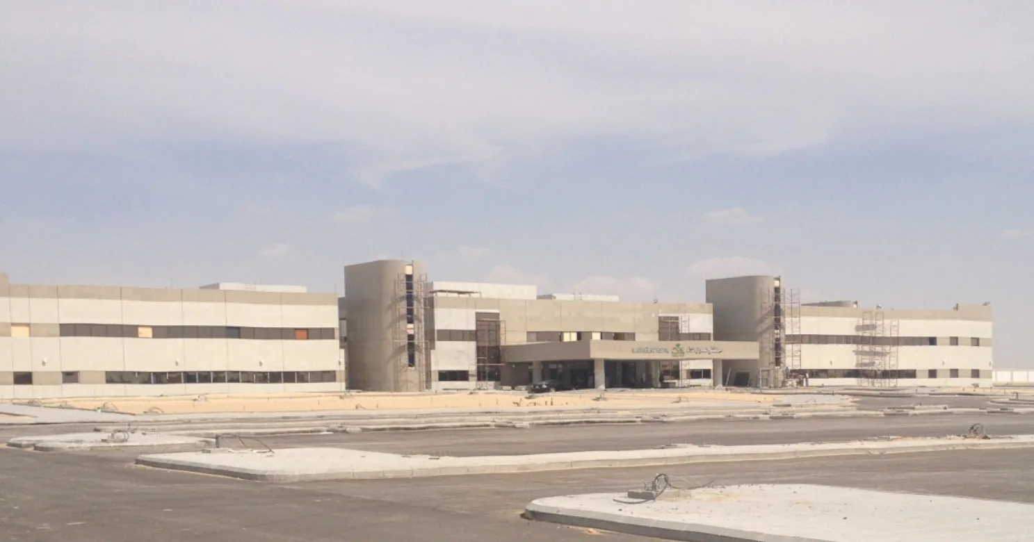 Hospital exterior in grey-beige cladding with scaffolding still standing at both towers, and unfinished asphalt and kerbs in front.