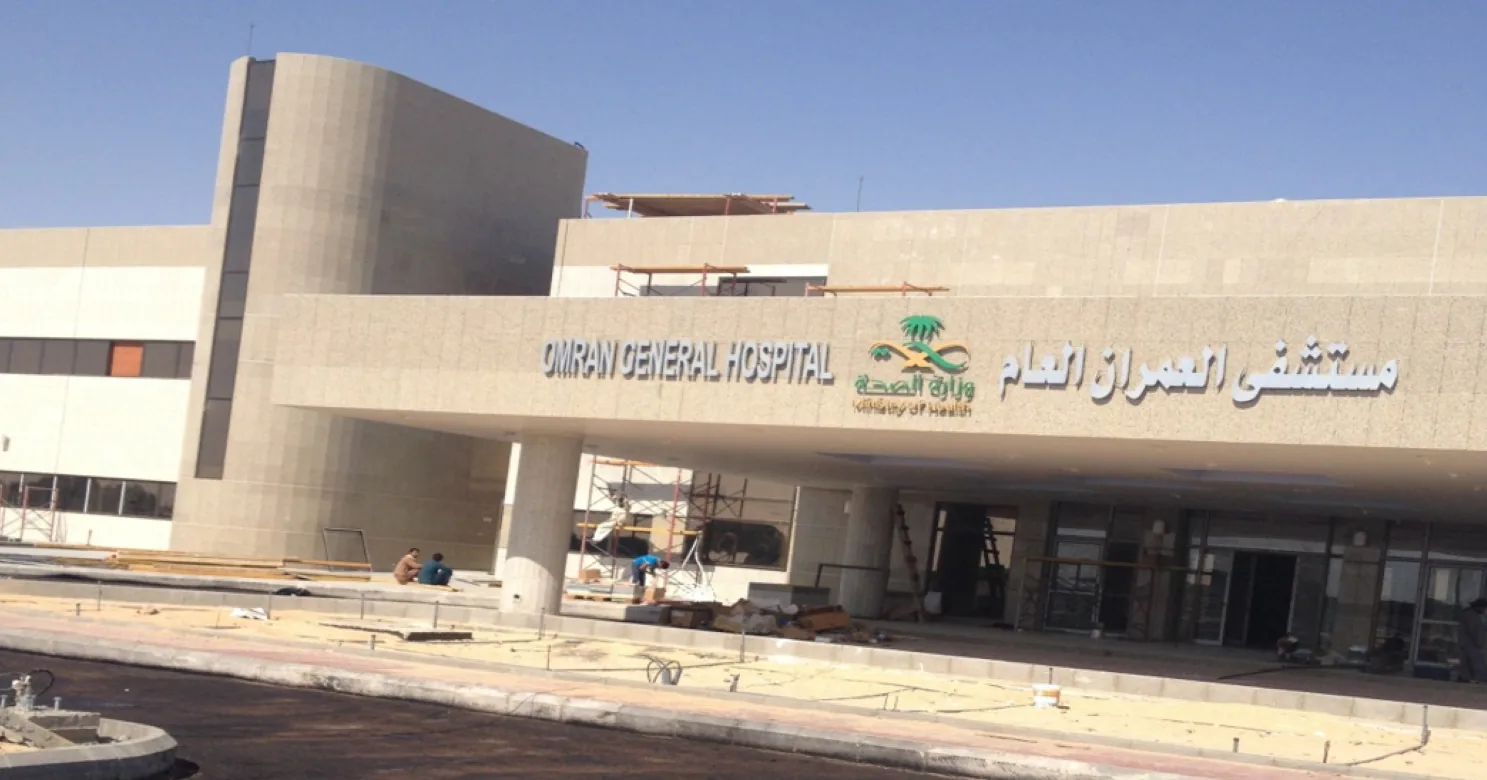 Hospital entrance canopy with English-Arabic name sign and Ministry of Health logo, scaffolding and workers at the doors, paving not yet laid.