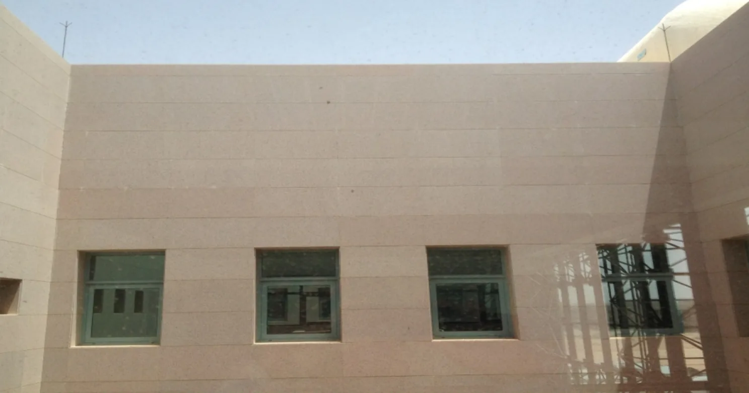 Beige stone cladding panels on a courtyard wall in horizontal courses, with three green-framed windows and scaffolding reflected at right.