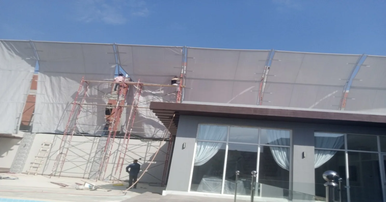 Workers on red scaffolding fitting fabric membrane to the steel-framed canopy above a glazed pavilion with curtained windows.