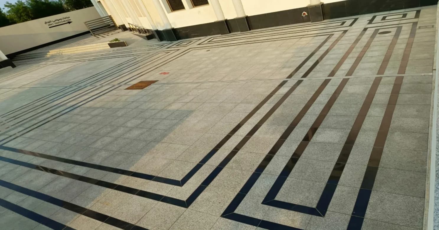 Wide finished granite courtyard in large slabs with multi-line black and brown inlay borders, steps and signage beside a white building.