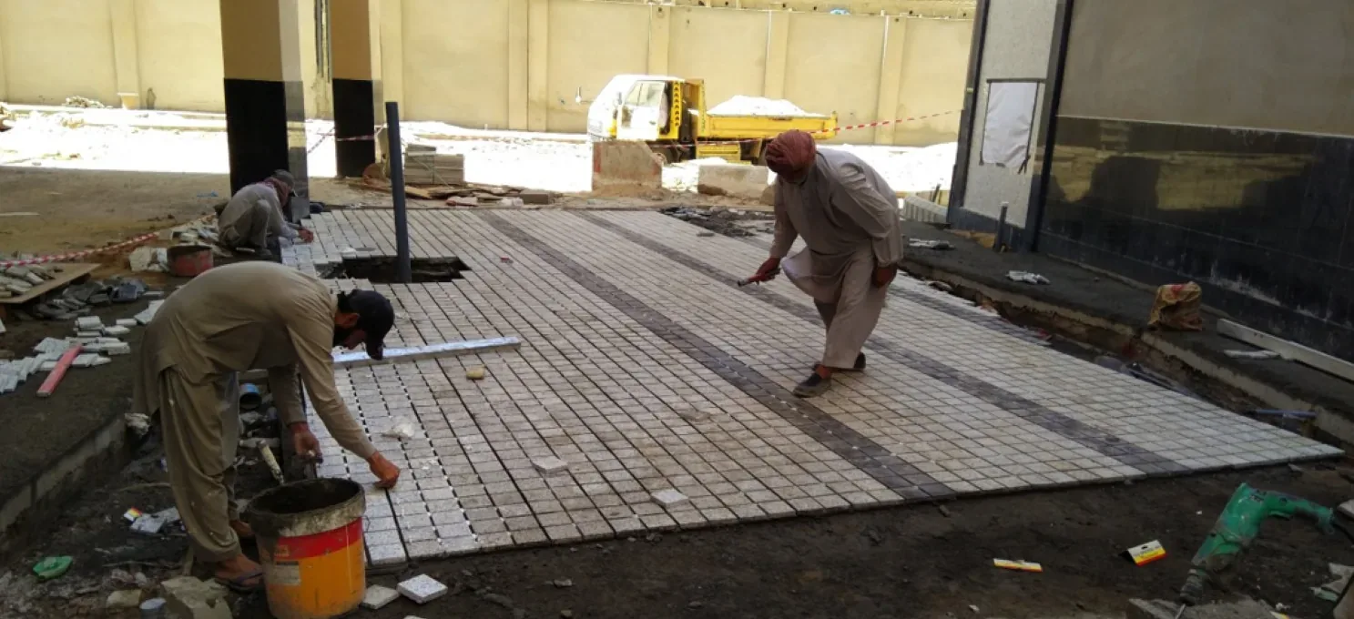 Two workers laying small-format granite mosaic with a dark band on a prepared bed, beside column bases, tools and an open drain.